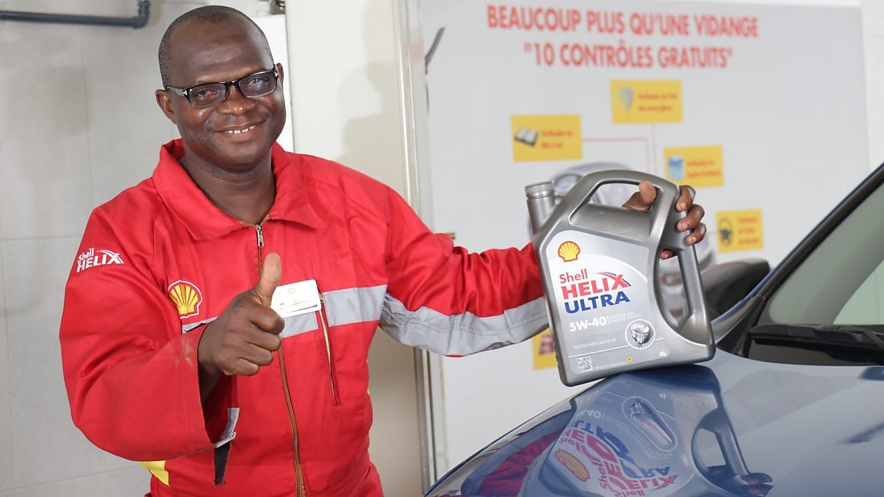 Technicien Shell tenant une bouteille d'huile Shell Helix devant un véhicule dans une station-service shell.