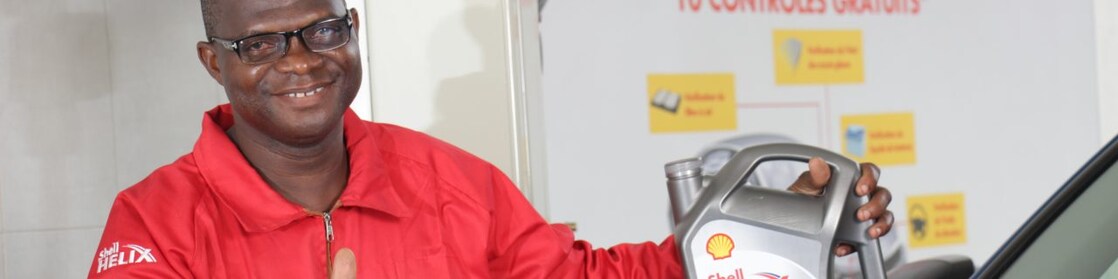 Technicien Shell tenant une bouteille d'huile Shell Helix devant un véhicule dans une station-service shell.