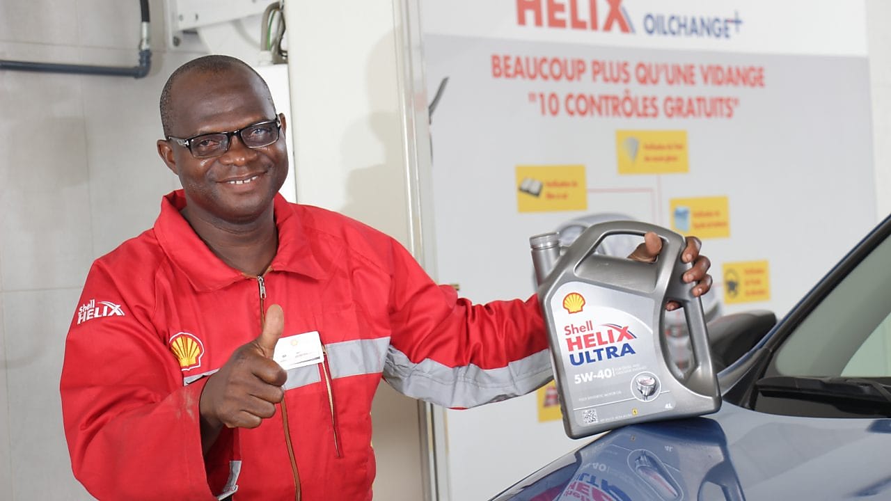 Technicien Shell tenant une bouteille d'huile Shell Helix devant un véhicule dans une station-service shell.